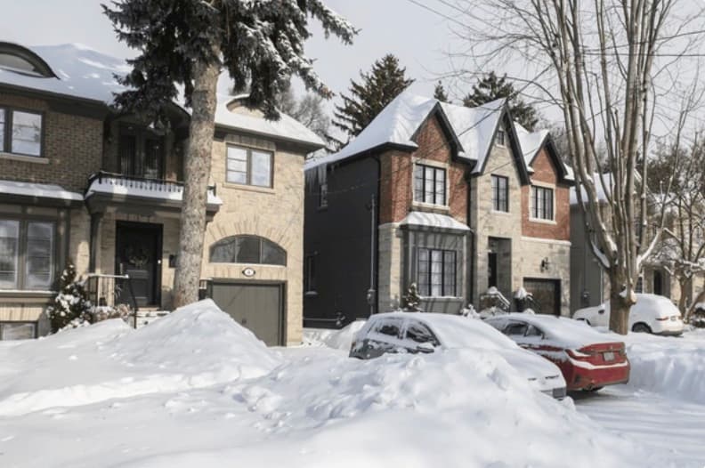 A snowy Toronto residential street: brick and stone homes with snow-covered roofs, cars buried under fresh snow along the curb