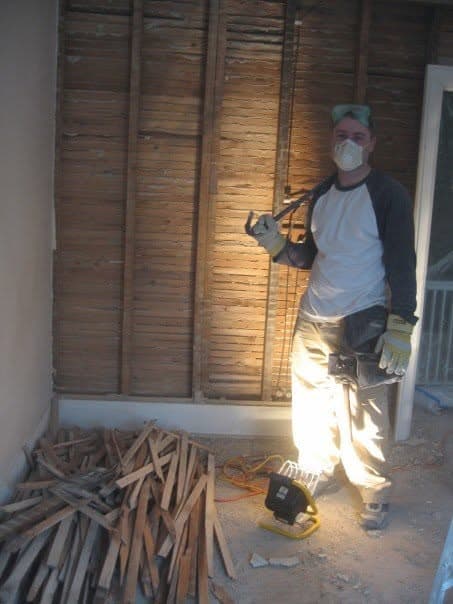 A person in a dust mask standing beside a wall of exposed wood lath, with a pile of removed lath on the floor mid-demolition