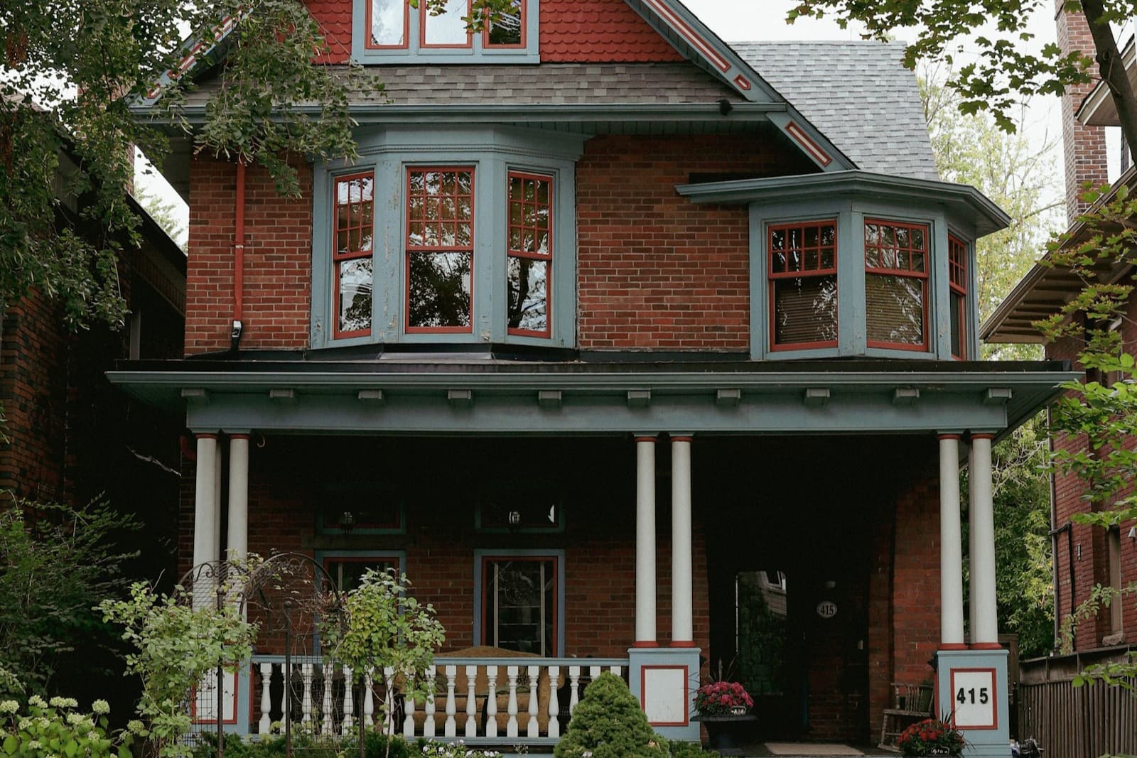 A red-brick Victorian Toronto home with blue-grey trim, bay windows, and a columned front porch on a leafy street