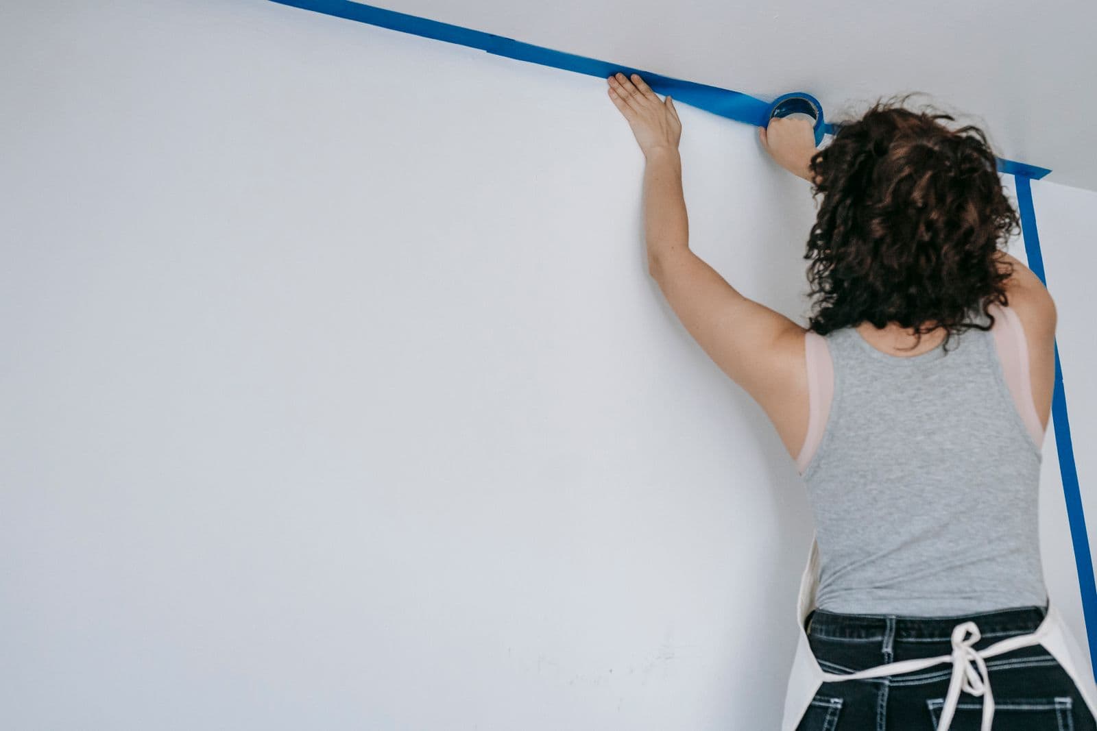 A painter applying blue painter's tape along the ceiling line of a bright white wall before painting